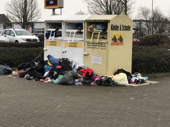 Blick auf drei Einwurf-Sammelcontainer (~1.5x1.5x2.5m) für Textilien/Kleider und Schuhe. In den Einwurfschlitzen liegt Kleidung, welche wegen Überfüllung offenbar nicht mehr rein passte. Davor und daneben liegen mehr als ein Dutzend große, teils aufgerissene Tüten mit weiteren Kleidungsstücken und Schuhe.
