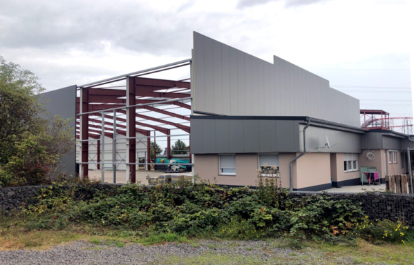 Rohbau einer Halle, geschätzt ~8-10m Höhe. Dunkelrote Stahlträger. Vorne, links hinten (74%) und rechts hinten (~50%) mit Metallplatten verkleidet. Vor der Halle direkt anschließend eingeschossiges Bestandsgebäude. Im Hintergrund Rutschen und Achterbahn zu erahnen.