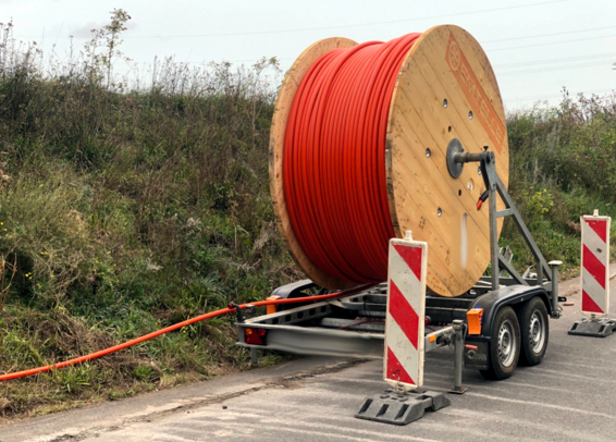 Abrollanhänger vor Böschung mit ~5m Durchmesser Rolle. Warnbarken vor und hinter dem Anhänger. ~85% mit orangem Plastik-Rohr bewickelt. Rohr geht nach hinten auf Böschung liegend ab. Sichtbare Frässpuren im Straßenbelag.