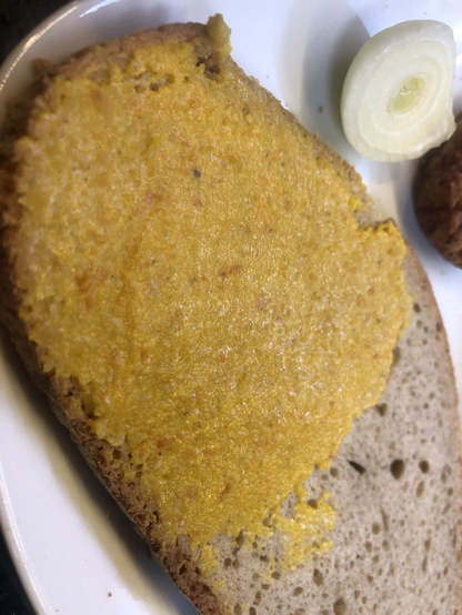 Brot mit orangem Muß. Kleine rot-orange Flecken erkennbar. Im Hintergrund eine angeschnittene Zwiebel und ein braunes etwas.