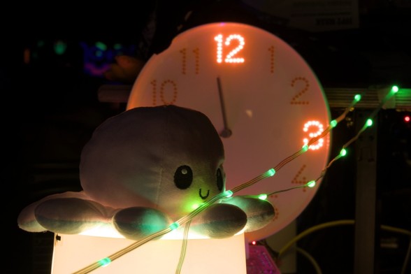Dunkle Szene
Vorne: Weißer, lachender Plüsch-Octopus auf eingeschalteter Tischlampe
Im Hintergrund: IKEA-Uhr, Ziffern durch LEDs gebaut, 12 und 3 leuchten.
Quer drüber: Lichterkette mit grünen Lampen.