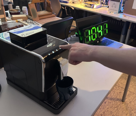 Blick auf einen Tisch. Links eine Kaffeemaschine mit Tasse, bei der Jemand grade eine Taste drückt.  Rechts davon eine grüne, 4-stellige 7-Segment-Anzeige mit der Zahl 1041