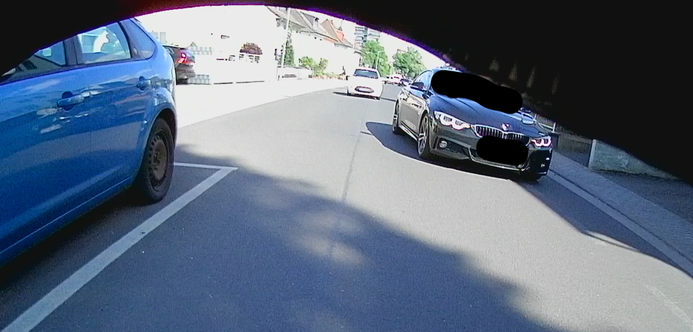 Rückansicht von einem Fahrrad. Links parkender PKW, etwas dahinter: Ein BMW, welcher in Richtung Hochbord unterwegs ist.