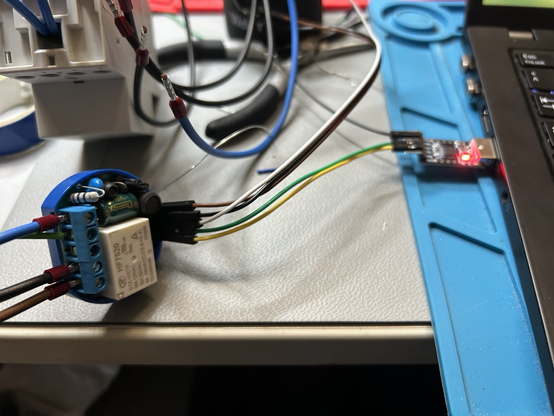 Geöffneter Shelly 1 mit Steckkabeln auf UART-Adapter am Laptop. An den Schraubterminals hängt ein nicht and Stromnetz angeschlossenes DIN-Gerät.