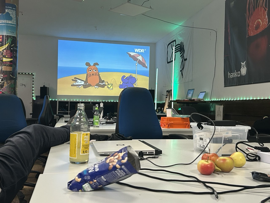 Blick in einen Hackspace um 03:30 morgens. Auf den Tischen ist allerlei Kabelkram, Laptops, Obst, Getränke, etc. Auf der Wand ist ein Beamerbild mit der Maus und dem Elefanten.