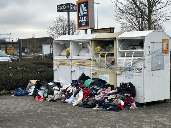 4 nebeneinanderstehende Kleidercontainer, welche mit Klarsichtfolie umwickelt wurden um diese zu sperren. Die Klarsichtfolie ist teilweise runtergerissen, in und vor den Containern türmen sich Berge von Textilien.