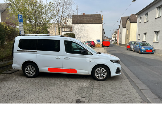 Blick von einem Fußweg. Links daneben Parkplätze. Auf einem Stellplatz steht ein Van, welcher neben der Parklücke auch den kompletten Fußweg blockiert. Einige Meter später parkt ein LKW auf dem Fußweg. Auf der anderen Straßenseite ist kein Fußweg angelegt, am Straßenrand parken weitere Fahrzeuge.
