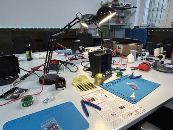 Tisch mit mehreren Lötplätzen. Silikonunterlage, Lötkolben, Lötzinn, Pinzetten und Zangen, Lampen, etc.