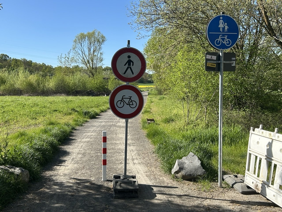 Ein Feldweg mit wassergebundener Oberfläche. Links Wiese, rechts Wiese mit Bäumen. Einfahrt rechts und links mit Felsen begrenzt. In der Mitte ein rot/weißer Pfosten. Rechts Mast mit Vz 240 (Gemeinsamer Geh- und Radweg). Mitten auf dem Weg ein mobiler Mast mit Vz 259 ("Verbot für Fußgänger") und 254 ("Verbot für Radfahrer")