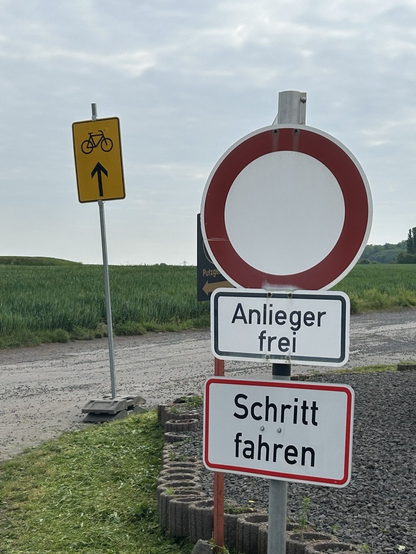 Fester Mast mit 250 (Verbot für Fahrzeuge aller Art) mit Zusätzen 1020-30 (Anlieger frei) und einem nicht genormten, weißen, rechteckigem Schild "Schritt fahren" mit rotem Rand. Links daneben ein temporärer Aufsteller mit orangem Schild mit Fahrradpiktogramm und Pfeil in die mit Verbot belegte Straße.