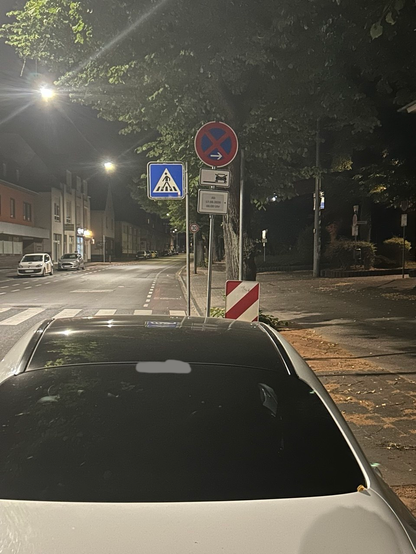 Straße bei Nacht. For der Kamera vvnh: Parkender BMW am rechten Fahrbahnrand, Schild Absolutes Halteverbot mit Zusatz auch a.d. Seitenstreifen und "Ab 17.08.2026 06:00", Fußgängerüberweg, Weiterer Straßenverlauf (30) mit Radschutzstreifen rechts.