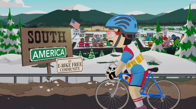Stadtansicht SouthPark/SouthAmerica. Ortsschild South, unterer Teil mit einem "America" überdeckt. Zusatzschild "an E-Bike-free community". Auf der Straße Randy Marsh in Rennradkleidung auf einem Bio-Bike.